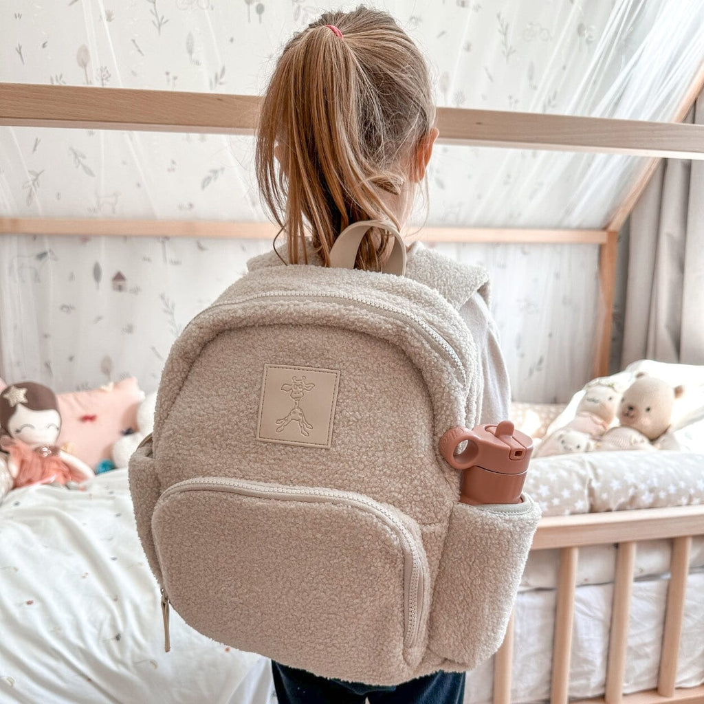 Little girl with ponytail carrying Ava Dusty Beige Mini Backpack on her back and using side pocket for her water/juice bottle.