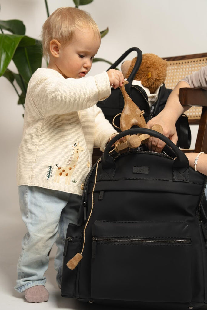 Mother holding open the main section of the Cost Changing Backpack while her small child places a wooden toy into the bag.