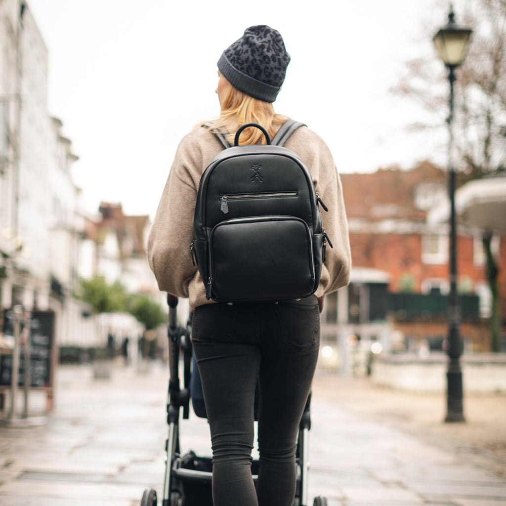a mum walking through a city in the UK, pushing a pram, with the black bloom changing backpack on her back