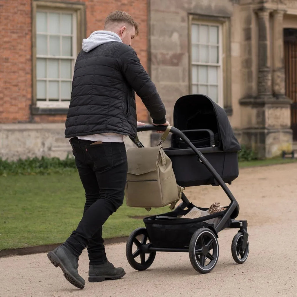 Dad pushing a pram through a country park with the luxe 2.0 changing backpack clipped onto the pram using the pram clips that come with the bag